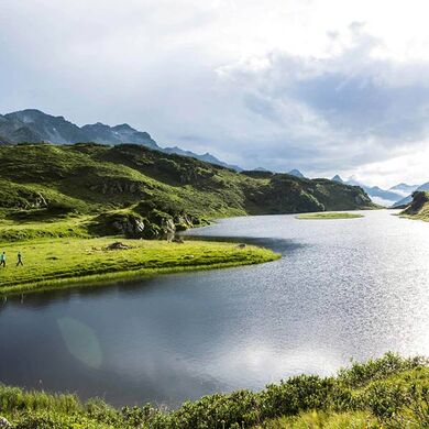 A long mountain lake in the Alps. Two people are walking along the shore and enjoying nature.