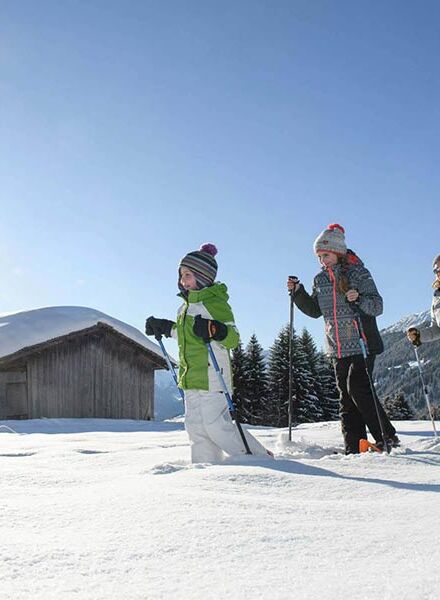 Eine Mutter geht mit ihrer Tochter und ihrem Sohn Schneeschuhwandern dabei gehen sie im Tiefschnee an einer Hütte vorbei