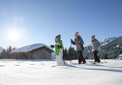 Eine Mutter geht mit ihrer Tochter und ihrem Sohn Schneeschuhwandern dabei gehen sie im Tiefschnee an einer Hütte vorbei