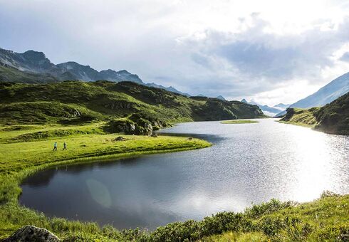 Ein langer Bergsee in den Alpen. Zwei Menschen gehen gerade am Ufer wandern und genießen die Natur.