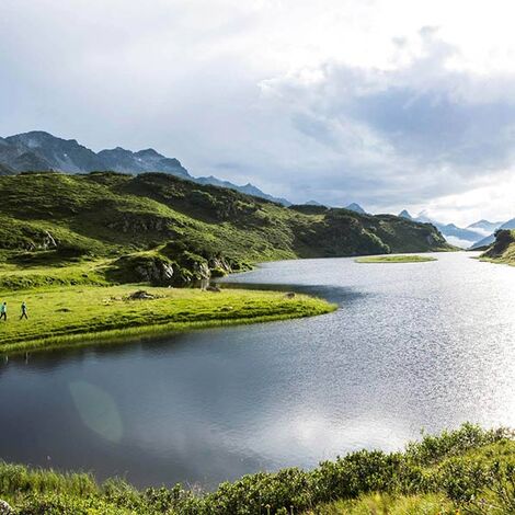 Ein langer Bergsee in den Alpen. Zwei Menschen gehen gerade am Ufer wandern und genießen die Natur.