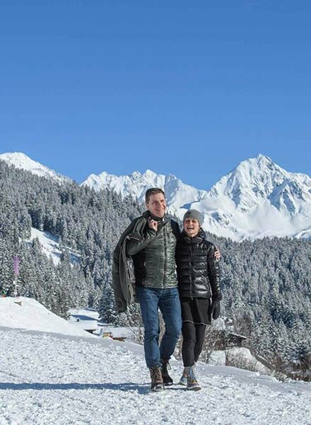 Ein Paar geht am Kristberg Wandern. Sie gehen Arm in Arm durch den Schnee und bestaunen das Winterpanorama.