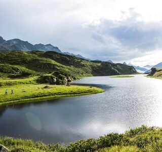 Ein langer Bergsee in den Alpen. Zwei Menschen gehen gerade am Ufer wandern und genießen die Natur.