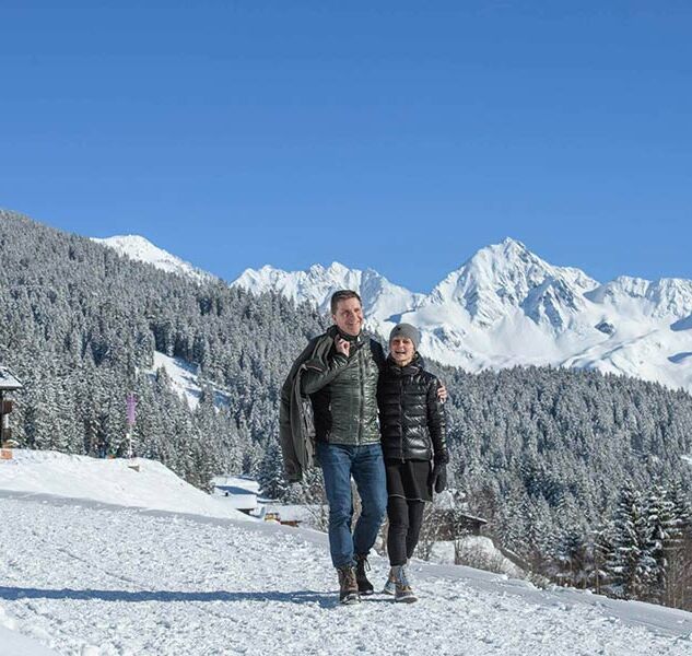 Ein Paar geht am Kristberg Wandern. Sie gehen Arm in Arm durch den Schnee und bestaunen das Winterpanorama.