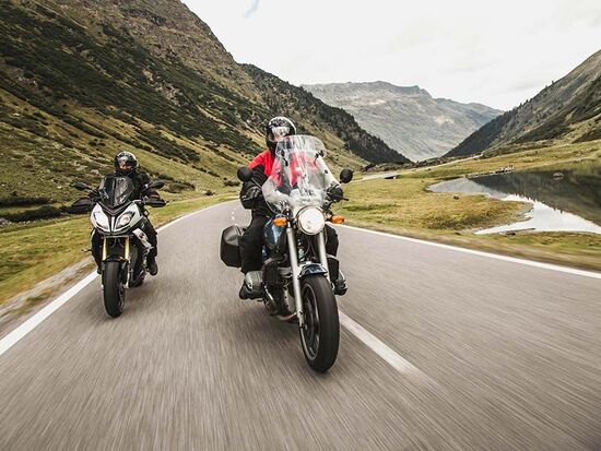 Two motorcyclists on the Silvretta High Alpine Road – a crystal-clear mountain lake in the foreground and the majestic peaks of the Silvretta mountains in the Austrian Alps in the background.