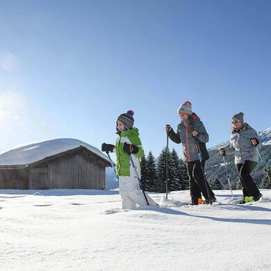 Eine Mutter geht mit ihrer Tochter und ihrem Sohn Schneeschuhwandern dabei gehen sie im Tiefschnee an einer Hütte vorbei