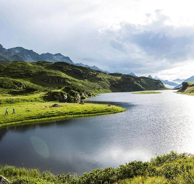 A long mountain lake in the Alps. Two people are walking along the shore and enjoying nature.