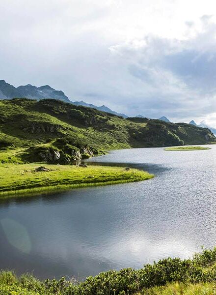 Ein langer Bergsee in den Alpen. Zwei Menschen gehen gerade am Ufer wandern und genießen die Natur.