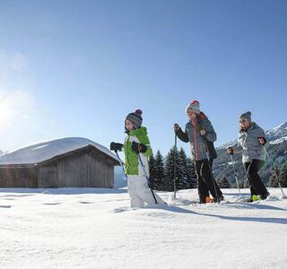 Eine Mutter geht mit ihrer Tochter und ihrem Sohn Schneeschuhwandern dabei gehen sie im Tiefschnee an einer Hütte vorbei