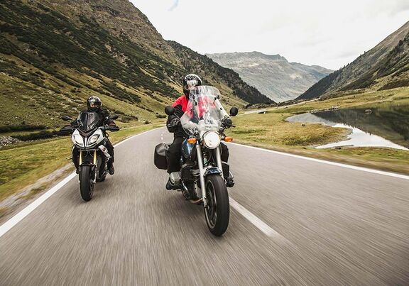 Two motorcyclists on the Silvretta High Alpine Road – a crystal-clear mountain lake in the foreground and the majestic peaks of the Silvretta mountains in the Austrian Alps in the background.