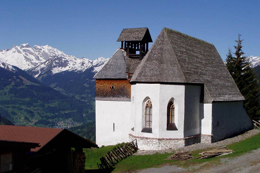 Frühlingsaufnahme der St. Agatha Kapelle am Kristberg im Silbertal, dem Genießerberg im Montafon in den österreichischen Alpen – im Hintergrund die verschneiten Drei Türme des Rätikongebirges.